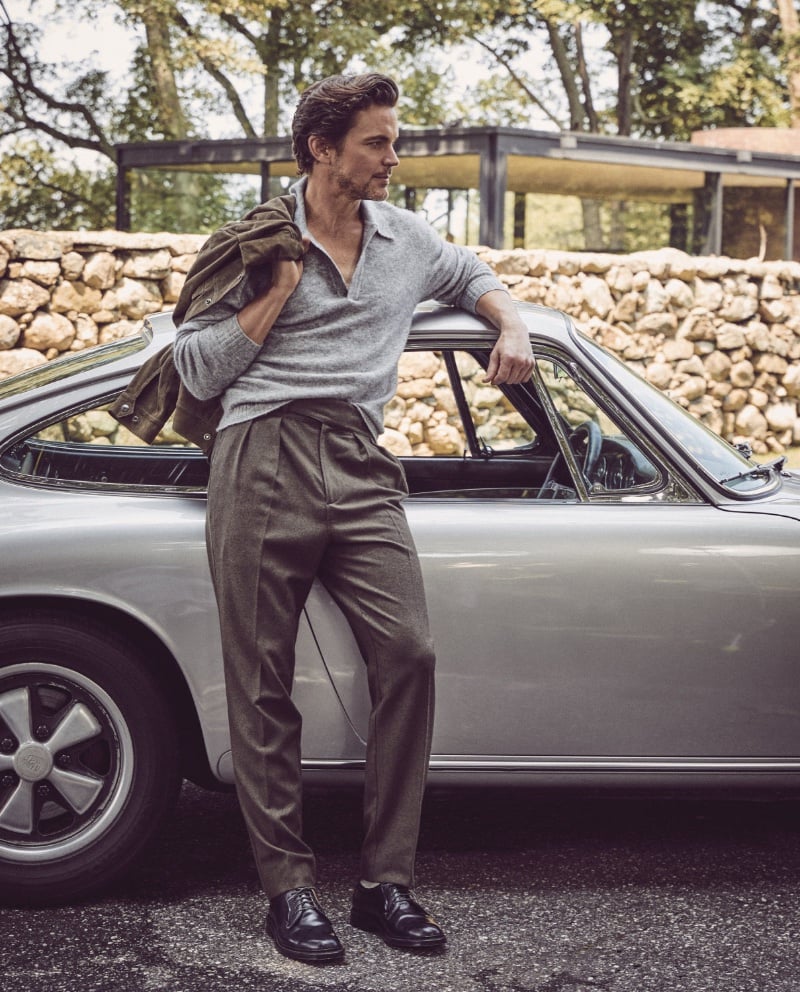 Matt Bomer leans against a classic car, sporting a sleek Todd Snyder alpaca popover polo, with sophisticated Gurkha trousers and Alden cordovan plain toe bluchers.