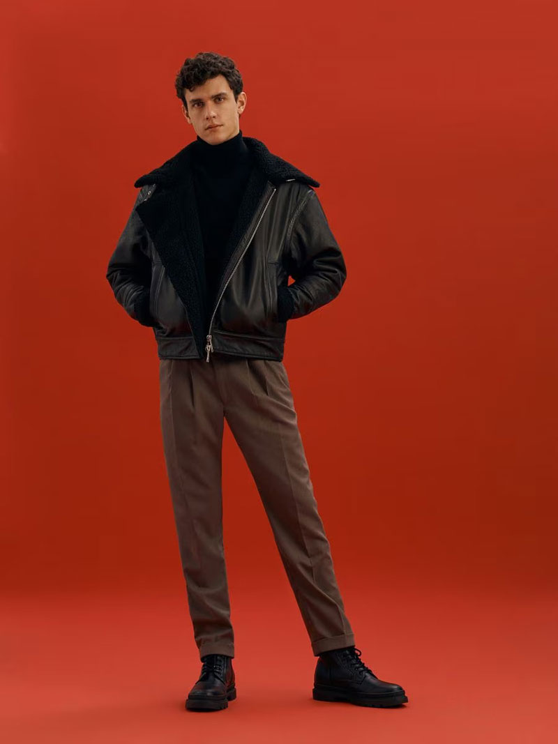Poised and stylish, Xavier Serrano stands against a vibrant red backdrop, sporting a chic black leather jacket over a turtleneck and tailored trousers.