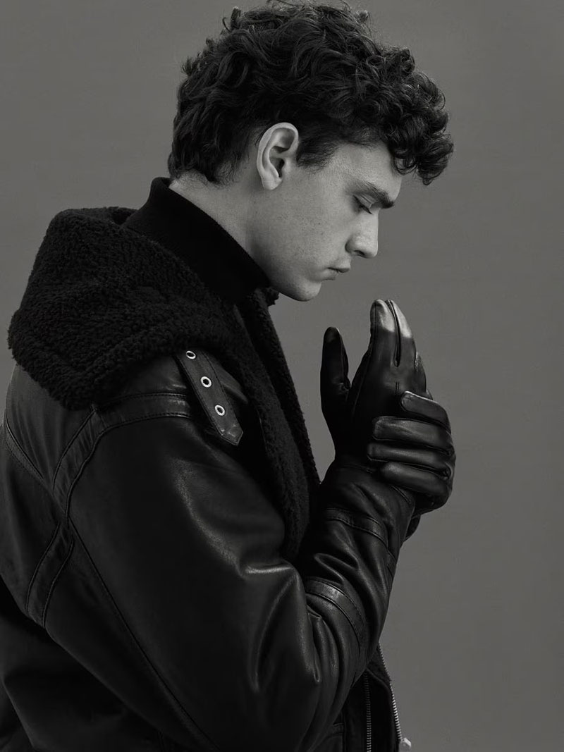A contemplative young man in a textured black leather jacket with fur collar, holding a thoughtful pose with eyes closed.