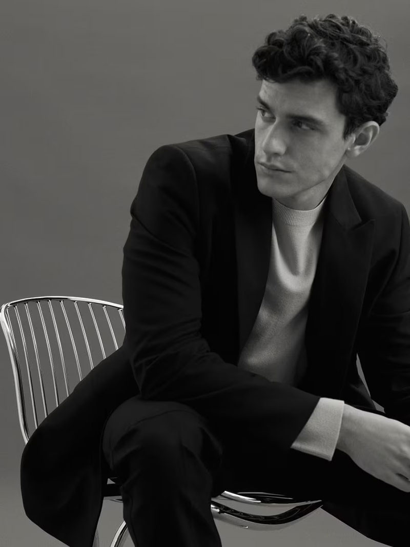 Model Xavier Serrano exudes elegance seated on a modern chair, dressed in a sleek black coat with a contrasting white turtleneck.