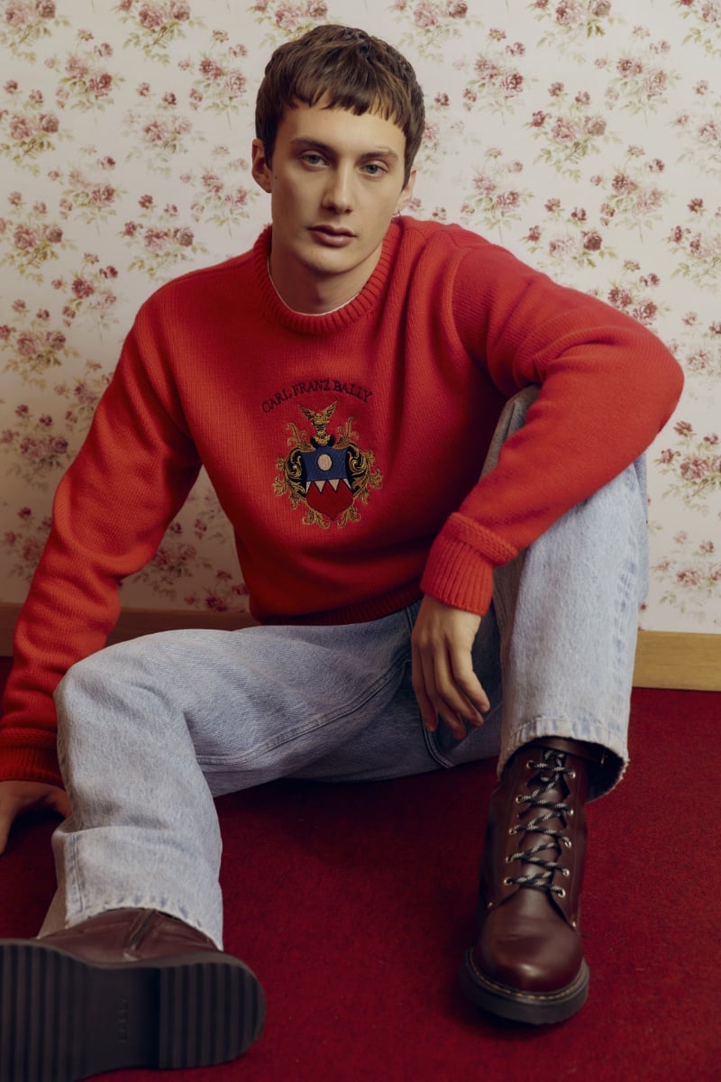 Gabriele Gratti brings a touch of vintage charm in a vibrant red sweater and relaxed light-wash jeans, grounded by sturdy brown boots, against a whimsical floral wallpaper for the Bally Mountain capsule collection.