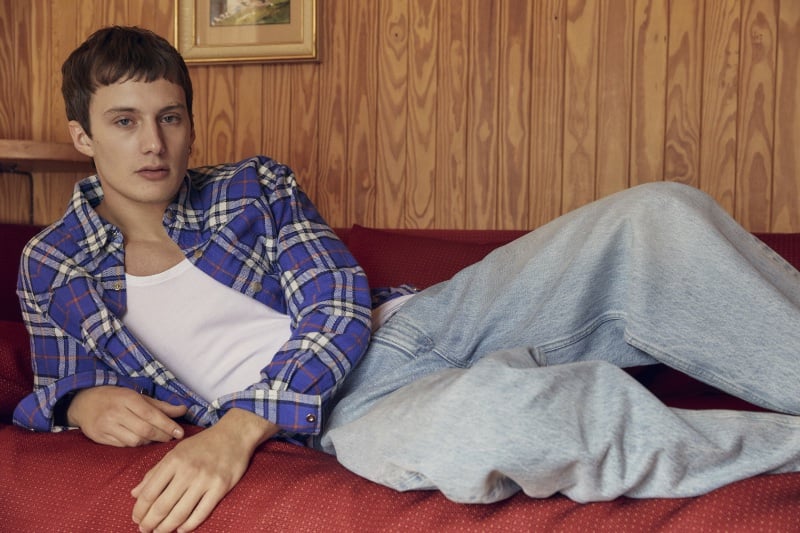 Gabriele Gratti lounges casually in a laid-back ensemble featuring a blue plaid shirt and light-wash jeans against the retro backdrop of a wood-paneled room. 
