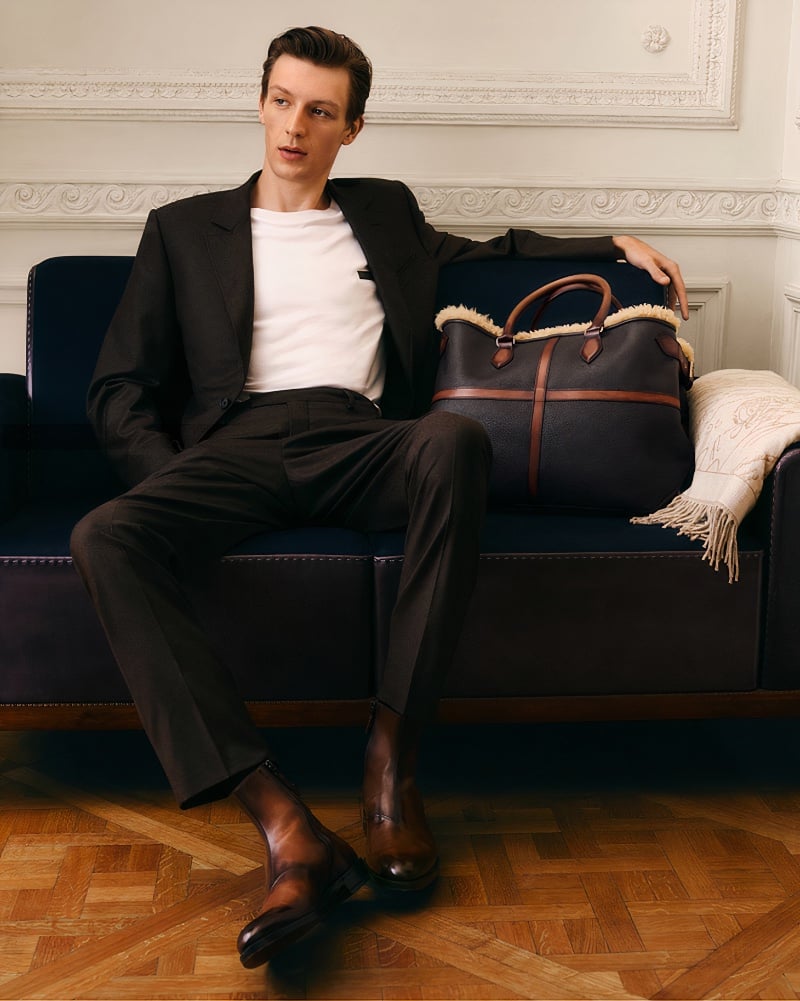 Relaxed yet refined, Finnlay Davis lounges, his style accentuated by Berluti's tailored trousers, polished leather boots, and a luxurious holdall.