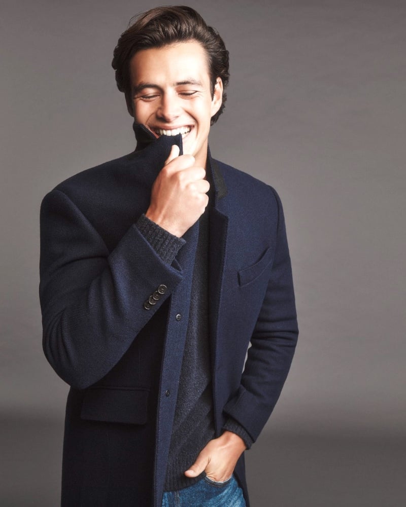 Harry Gozzett flashes a charming smile wrapped in a cozy navy coat layered over a textured sweater from Club Monaco. 