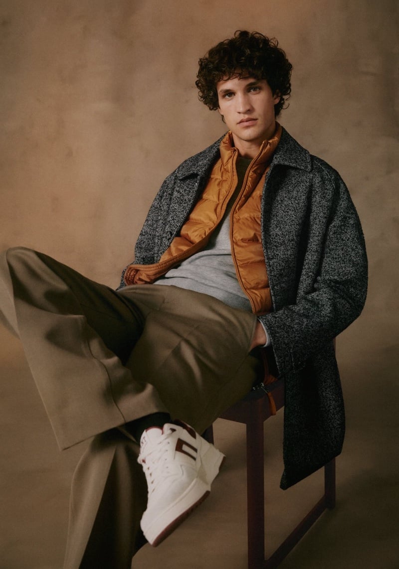 Model Francisco Henriques is the epitome of casual cool, seated in a salt-and-pepper GANT overcoat, puffer, and pleated trousers. 