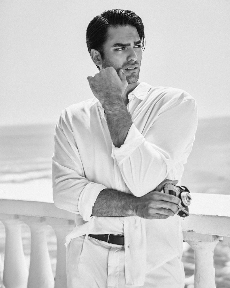 Matteo Bocelli exudes a thoughtful charm, holding a vintage camera while dressed in crisp white shirt, reflecting timeless seaside elegance for the GUESS holiday 2023 campaign.