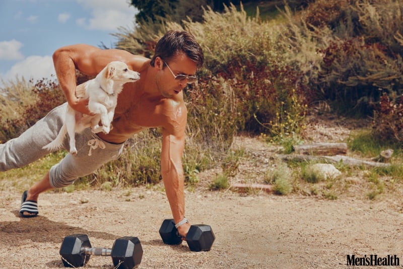 Glen Powell performs a one-armed push-up outdoors while balancing his dog Brisket. He wears Persol sunglasses with Todd Snyder x Champion sweatpants and an OMEGA watch.