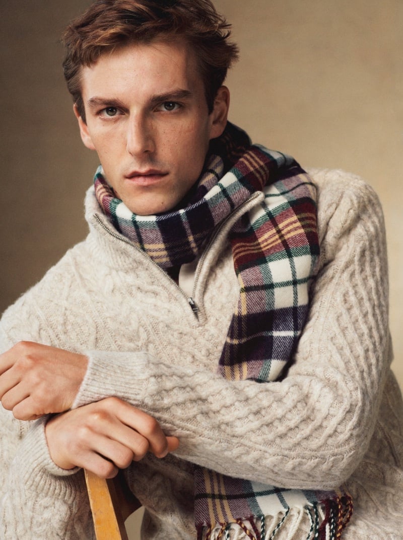 Model Quentin Demeester looks intently at the camera, wrapped in a cozy 3/4 zip cable knit sweater and a colorful plaid scarf.