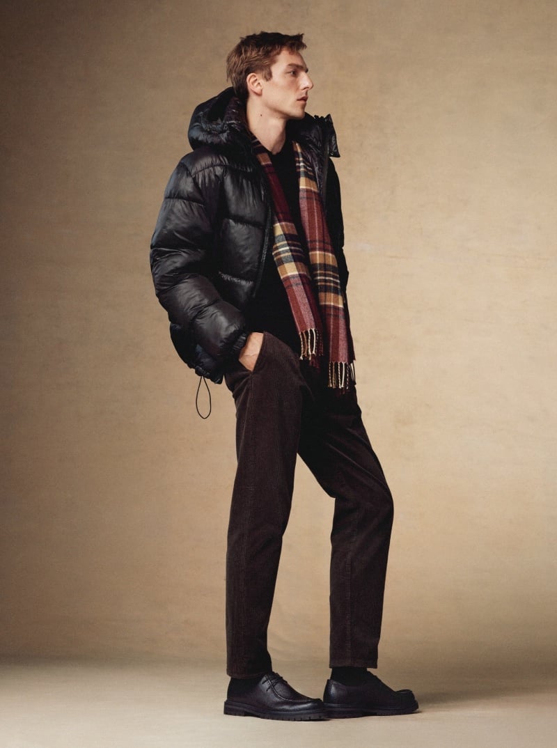 Quentin Demeester stands in profile, exuding a winter-ready vibe in a black puffer jacket accented by a classic plaid scarf and dark corduroy trousers. 