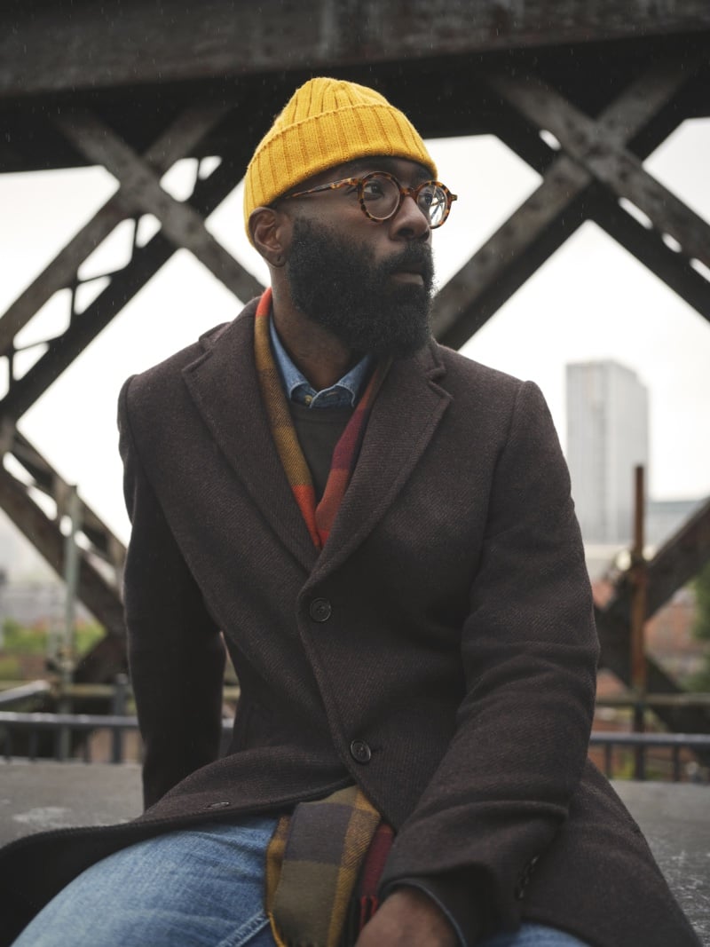 Paul Binam brings a pop of color to a cloudy day, layering a mustard beanie and a checkered scarf with a structured brown coat, embodying the casual yet refined spirit of the M&S Fall-Winter 2023 campaign.