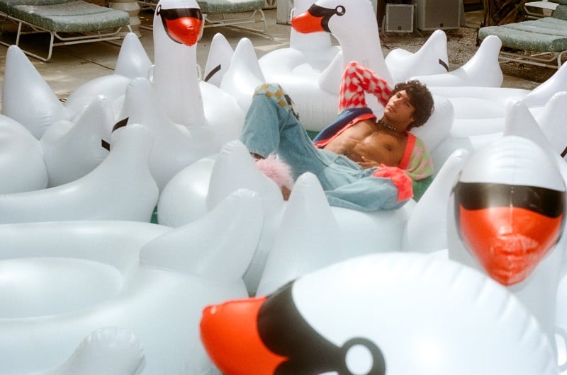 Carlos Mata relaxes on an inflatable swan, wearing a Marni multicolor cardigan and blue jeans for SSENSE.