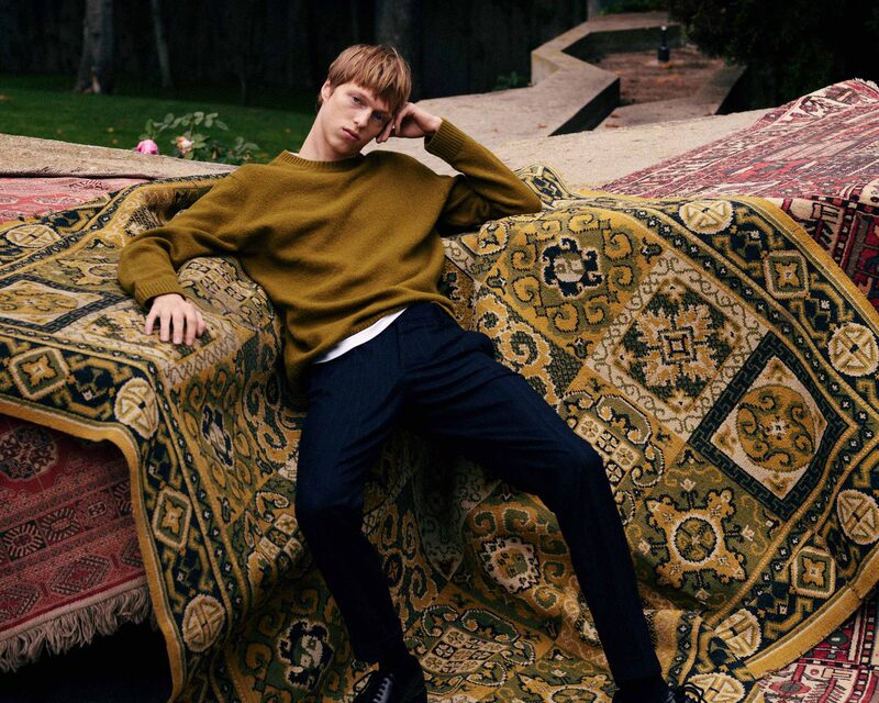 Model Joseph Uyttenhove lounges against a tapestry of ornate rugs, donning a mustard sweater and navy trousers.