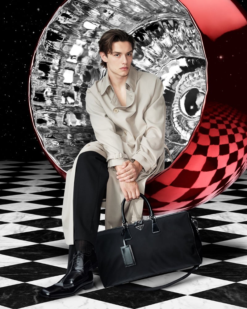 Donning a trench, Louis Partridge is seated against a surreal checkerboard backdrop for Prada's holiday 2023 campaign.