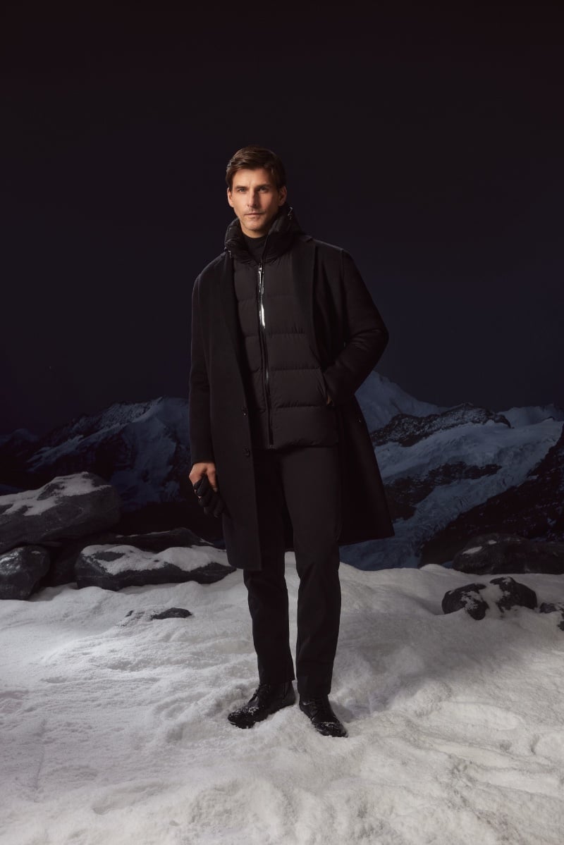 Johannes Huebl wears a black RUDSAK coat against the dramatic backdrop of a snowy mountain landscape.