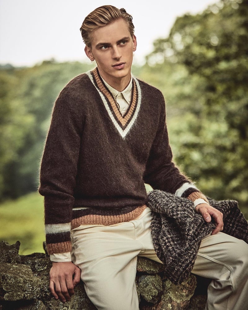 Dominik Sadoch sits atop a stone wall, sporting a rustic wool v-neck sweater from Todd Snyder.