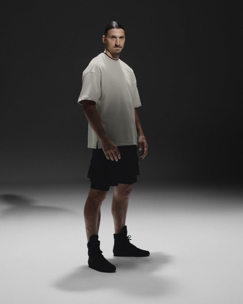 Zlatan Ibrahimović wears a casual H&M Move sportswear ensemble featuring a loose-fitting t-shirt and black shorts against a dimly lit backdrop. 