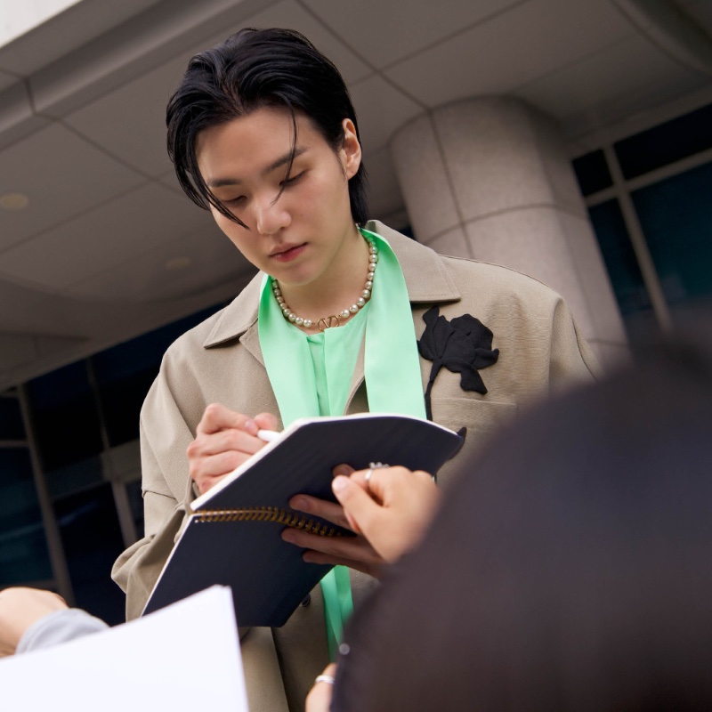 Suga signs autographs in The Narratives campaign, wearing a beige and green Valentino outfit.