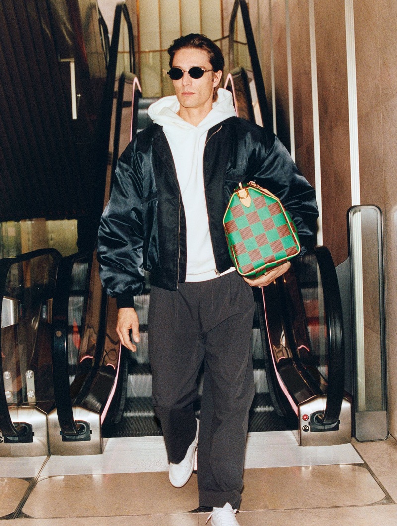 On the move, Francesco Mariottini makes an escalator descent, styled in an EZR jacket, Rhude hoodie, and Saint Laurent sunglasses, with a Louis Vuitton bag in tow. 