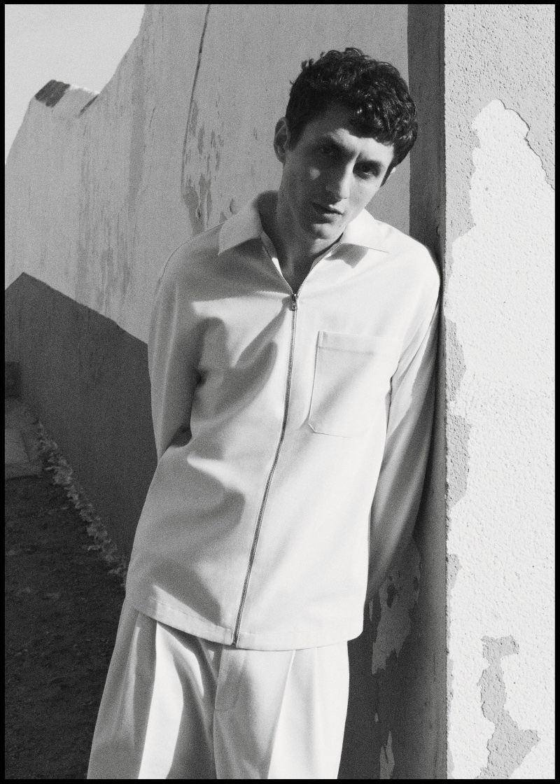 Henry Kitcher leans against an aged wall in a sleek white zip-up jacket from Mango.