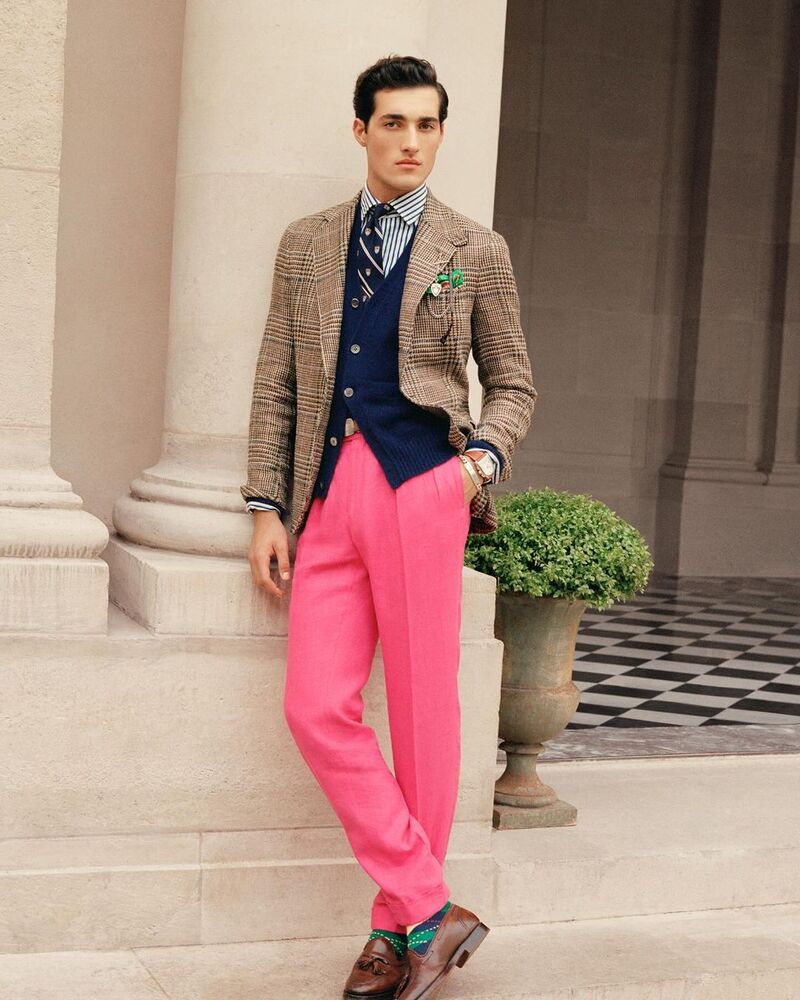 Adam Sattrup wears a Houndstooth blazer with a navy cardigan, striped dress shirt, and pink pleated trousers. 