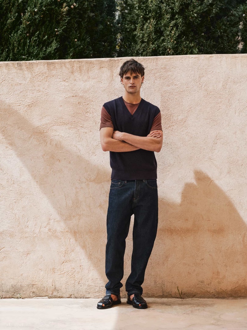 Showcasing Massimo Dutti's relaxed style, Parker van Noord wears a sweater vest with a t-shirt, jeans, and leather sandals. 