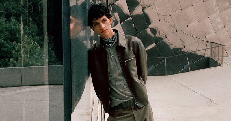 A model wears an olive leather jacket over a collared shirt and striped high-neck layer beside reflective architecture.