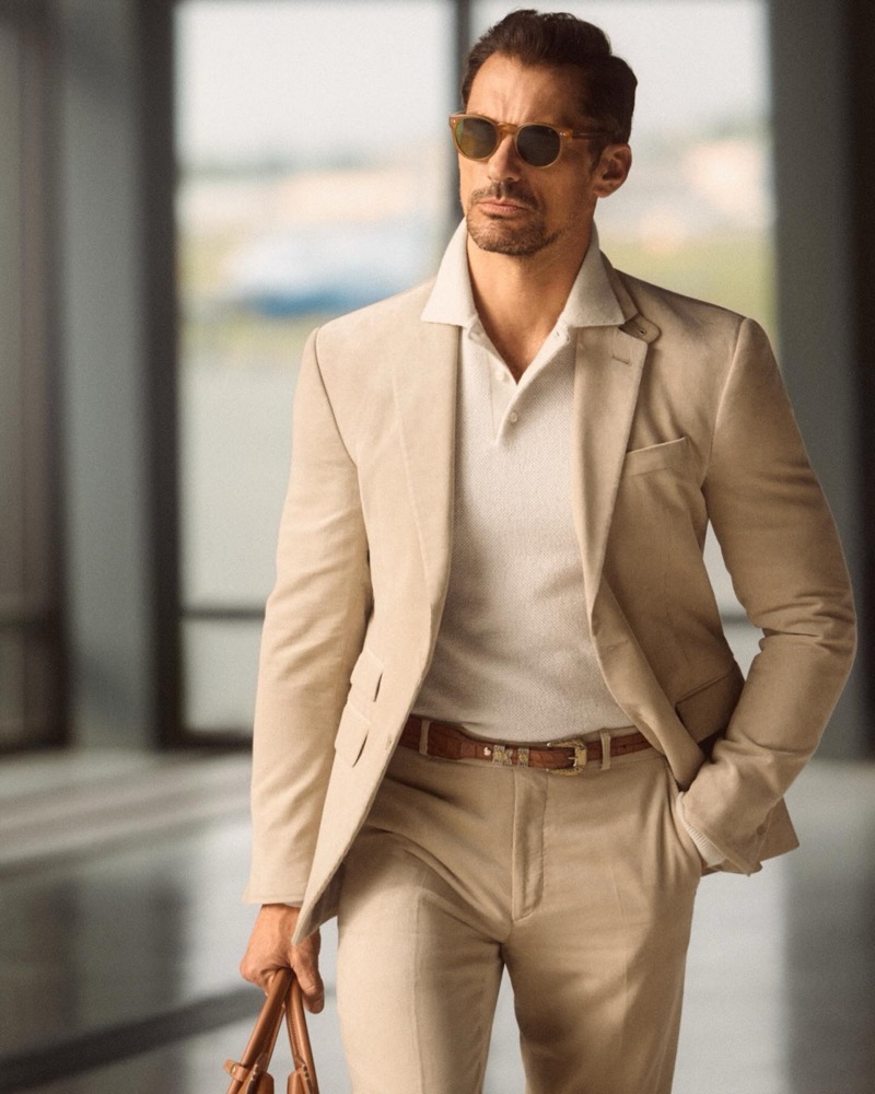 David wears a corduroy suit jacket with pleated trousers, a polo shirt and brown translucent framed sunglasses.