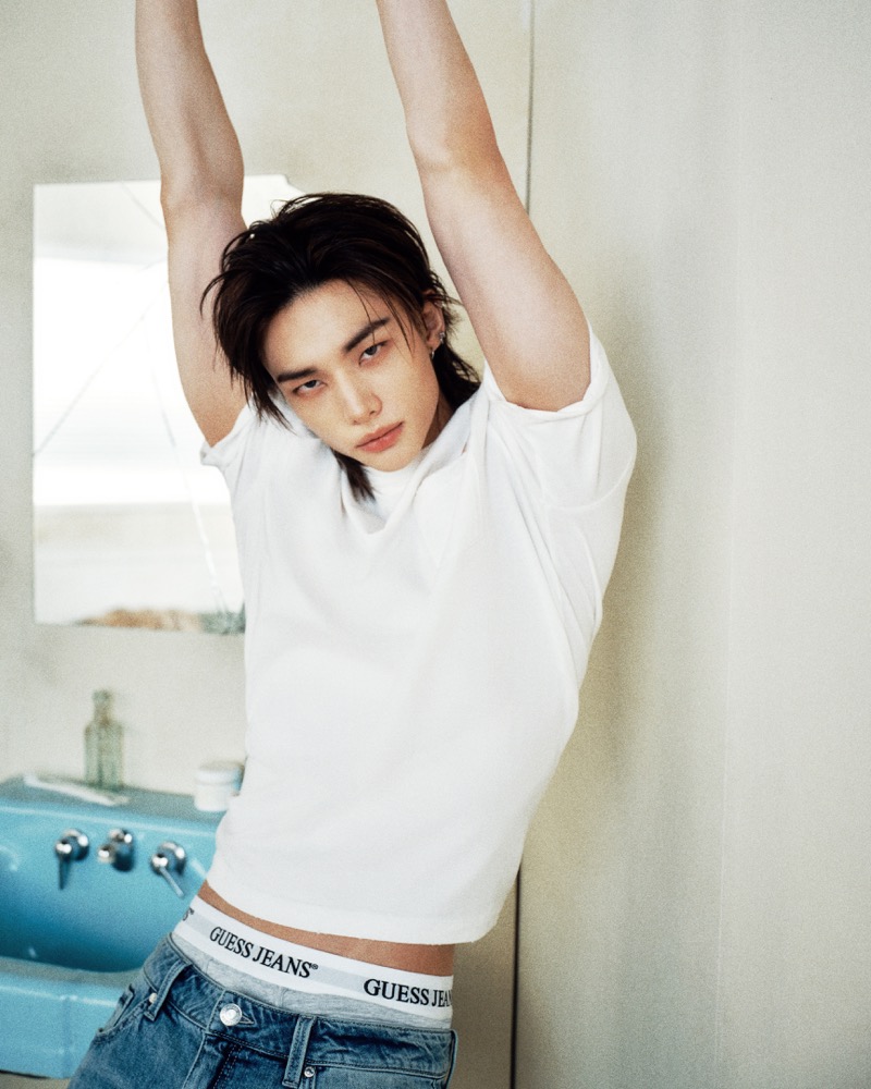 Hyunjin poses with his arms stretched above his head, wearing a white T-shirt, jeans, and GUESS Jeans underwear