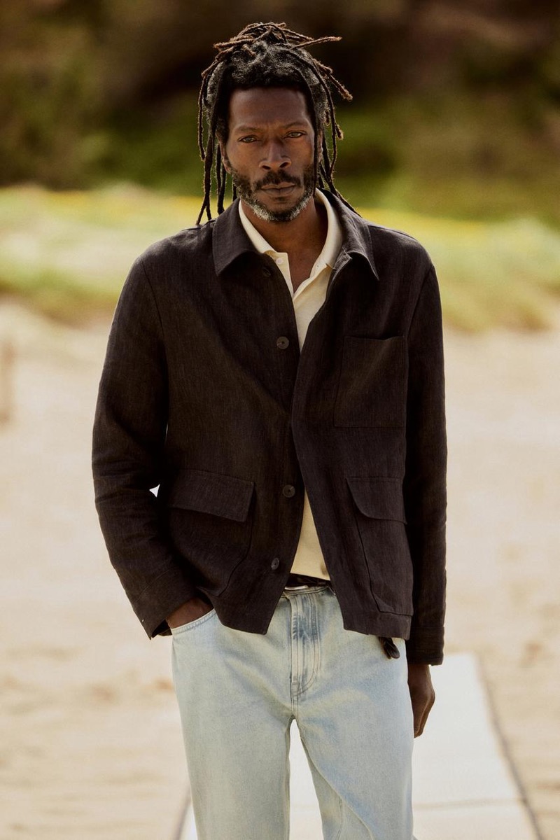 Virgil walks along the beach in a brown linen jacket with light wash jeans