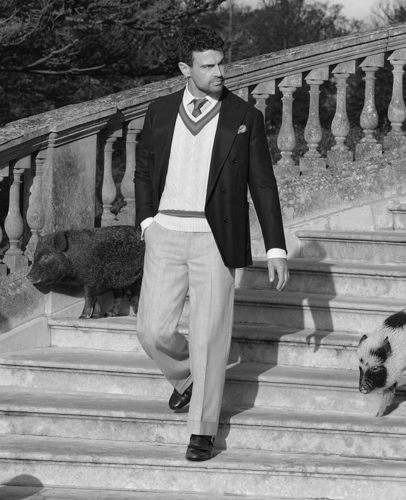 Theo descends a stone manor staircase in a double-breasted blazer, V-neck cricket sweater, and pleated trousers alongside two pot-bellied pigs