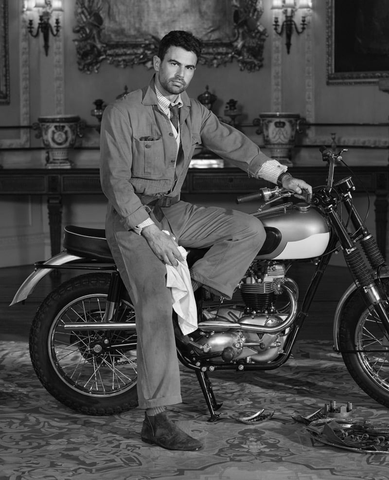 Theo seated on a vintage motorcycle inside a grand ornate manor room wearing a heritage work jacket and Reigate suede ankle boots