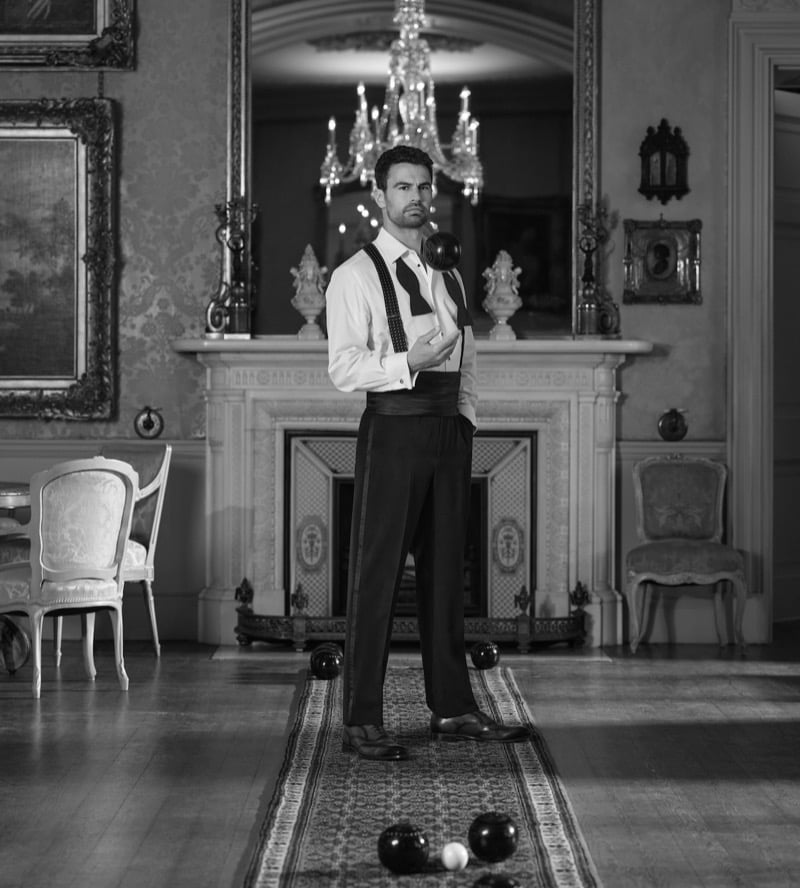 Theo James stands in a gilded manor interior in a white dress shirt, high-waisted black trousers, and suspenders with lawn bowls at his feet