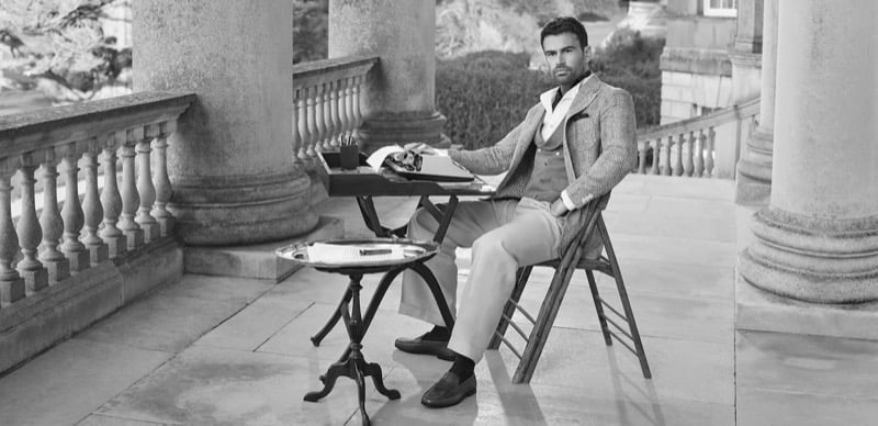 Theo sits at a typewriter on a stone colonnaded manor terrace in a linen suit jacket and Rother loafers