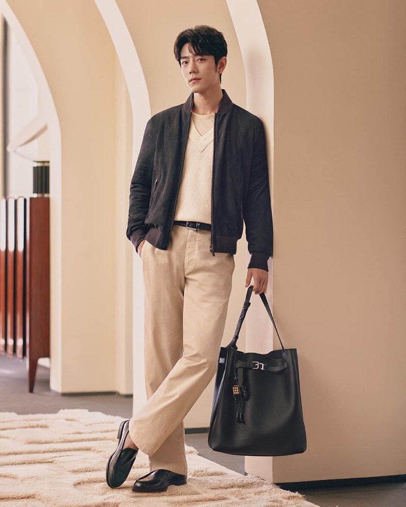 Xiao stands in an arched corridor wearing a dark suede bomber jacket, cream V-neck sweater, and khaki trousers while holding a black leather hobo bag