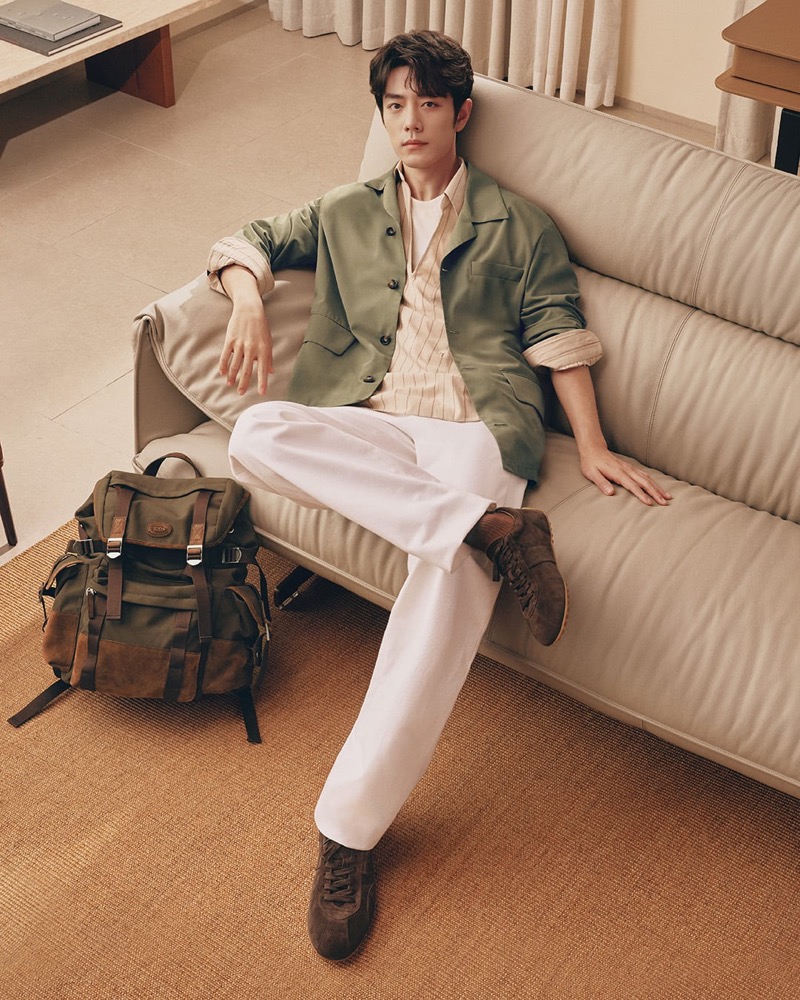 Xiao sits on a sofa in a green overshirt over a patterned popover and white trousers with brown suede sneakers