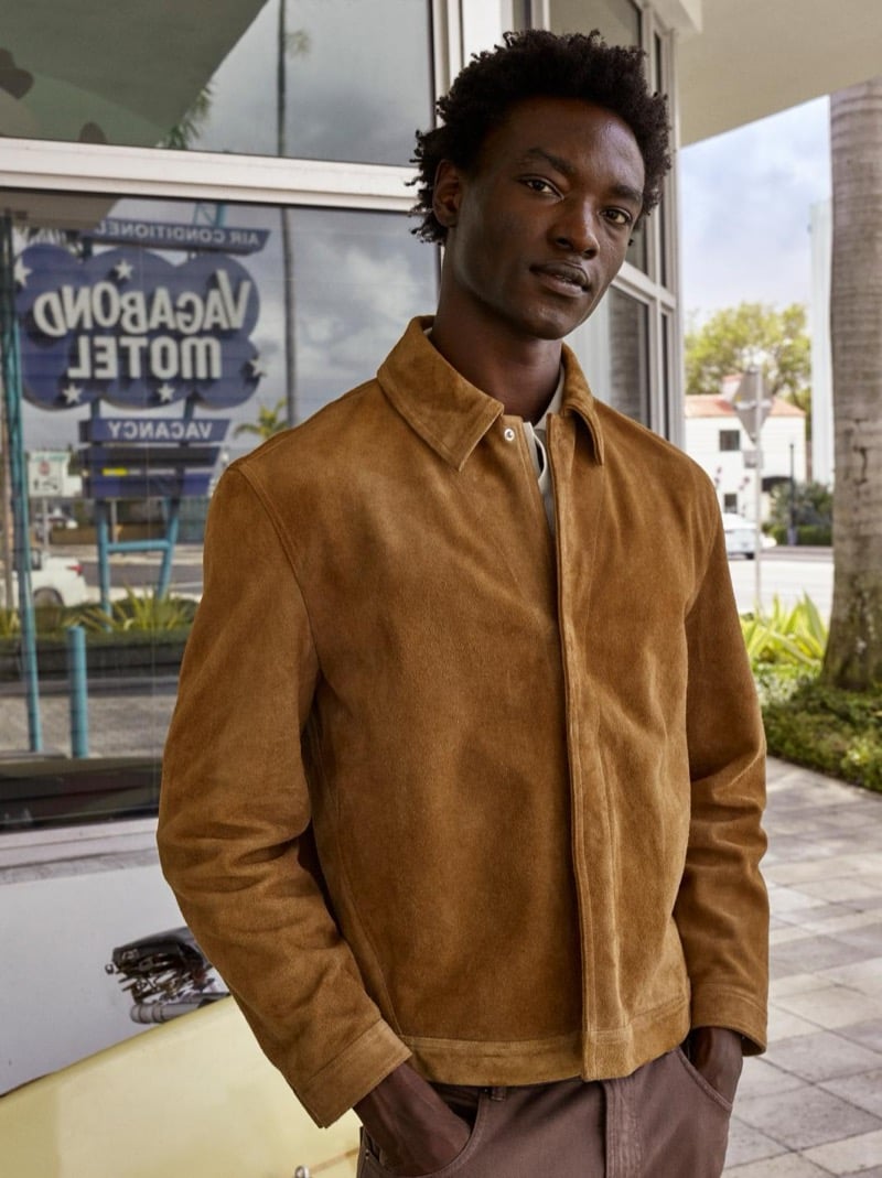George stands outside the Vagabond Motel in a tan suede overshirt and brown trousers