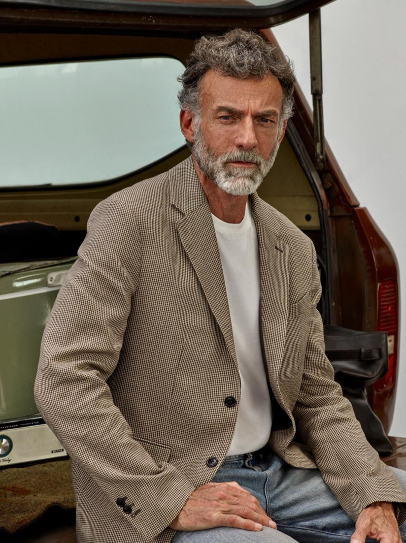 Alain sits at the boot of a vintage car in a houndstooth sport coat over a white crew neck and light-wash jeans
