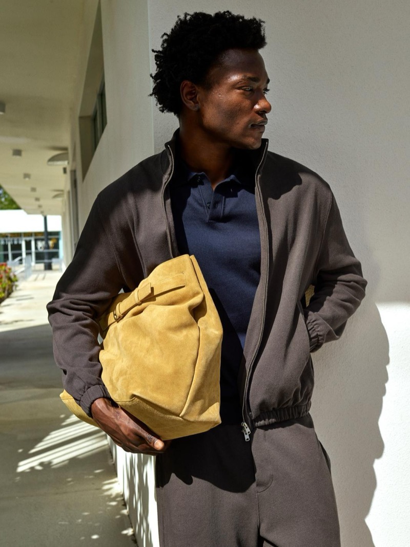 George wears a dark charcoal track jacket over a navy polo and holds a tan suede bag