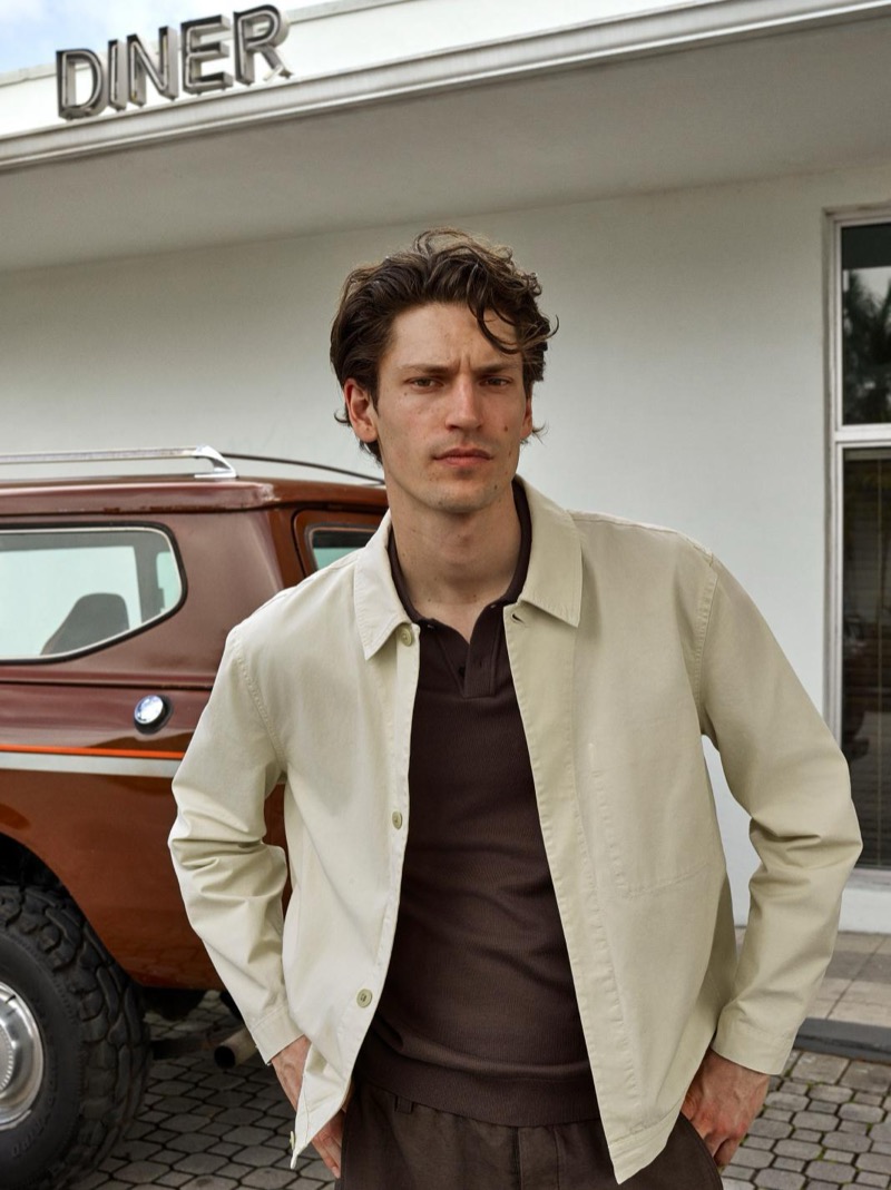 Justin wears a cream cotton overshirt over a dark polo and brown trousers outside a roadside diner