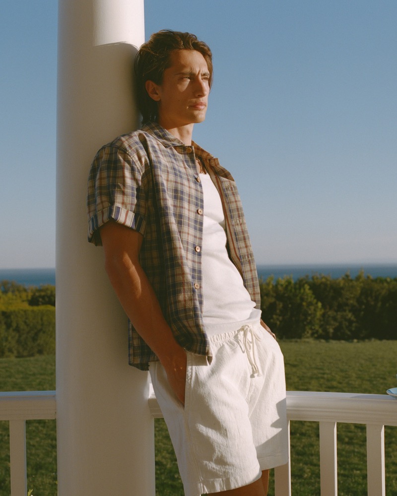James leans against a porch column wearing a plaid short-sleeve shirt open over a white tank and pull-on shorts