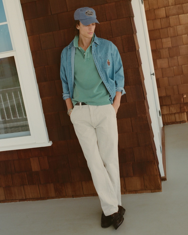 James leans against a shingled wall wearing a denim shirt over a green polo and striped relaxed trousers with a cap