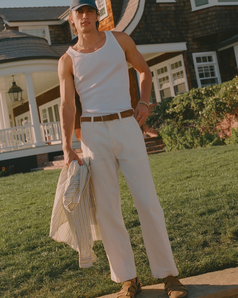 James stands on a lawn in a white tank top and cream relaxed trousers with a tan belt and suede sandals