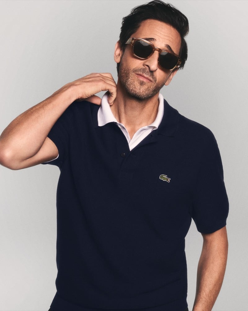 Brody wears a navy Lacoste polo shirt with a white contrast collar and caramel translucent acetate sunglasses with dark tinted lenses, one hand raised near his shoulder for Lacoste's spring 2026 eyewear campaign