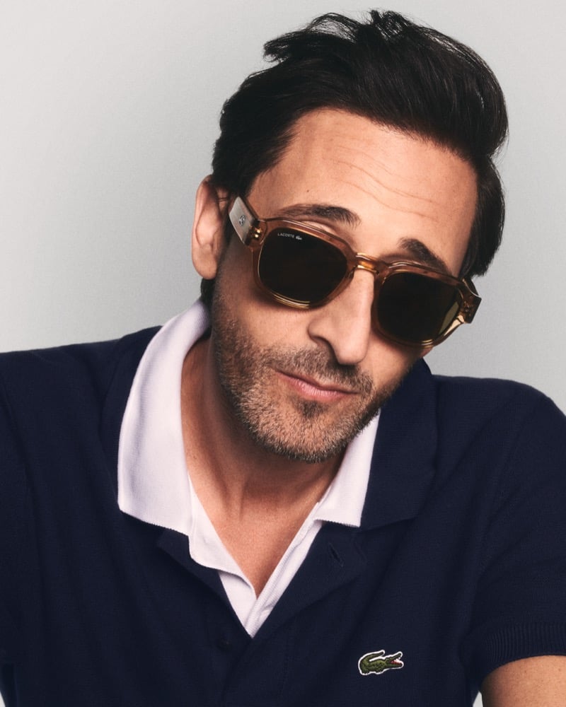 Close-up of Brody in caramel translucent acetate sunglasses with dark lenses and a visible Lacoste logo on the temple, wearing a navy polo with a white contrast collar for Lacoste's spring 2026 eyewear campaign