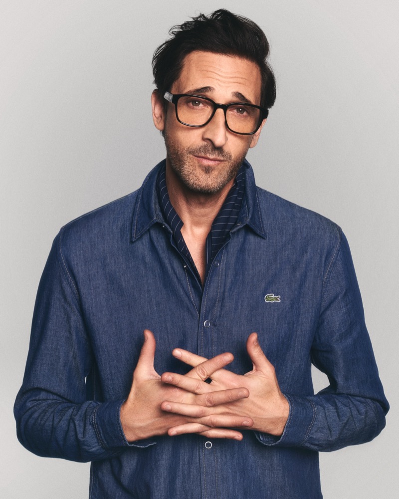 Brody stands with hands folded at his chest in a denim button-up shirt layered over a navy pinstripe shirt, wearing black square-frame optical glasses for Lacoste's spring 2026 eyewear campaign