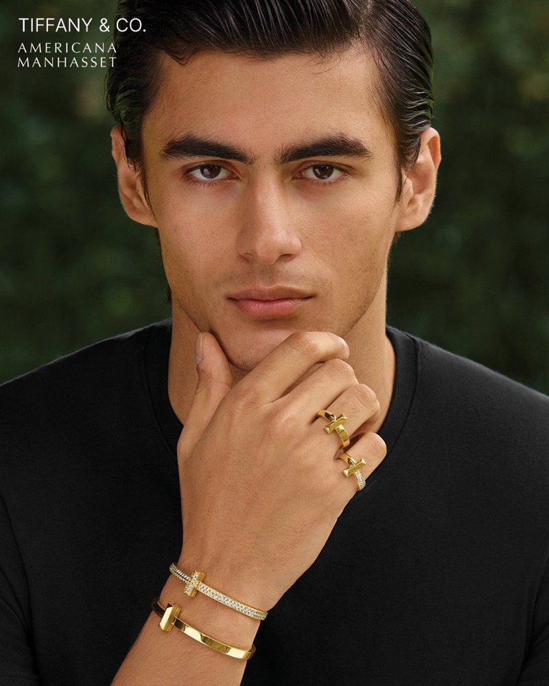 Akbar wears Tiffany & Co. gold T rings and a bracelet with a black tee in a close-up portrait