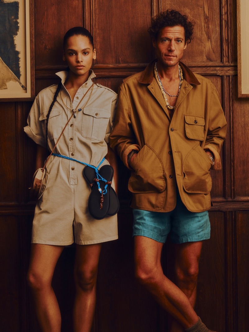 A female model wears a beige short-sleeve safari-style jumpsuit with a black crossbody flap bag, while Phillips wears a walking jacket with corduroy collar, turquoise shorts, and a beaded necklace for Banana Republic's Archive Reissue collection