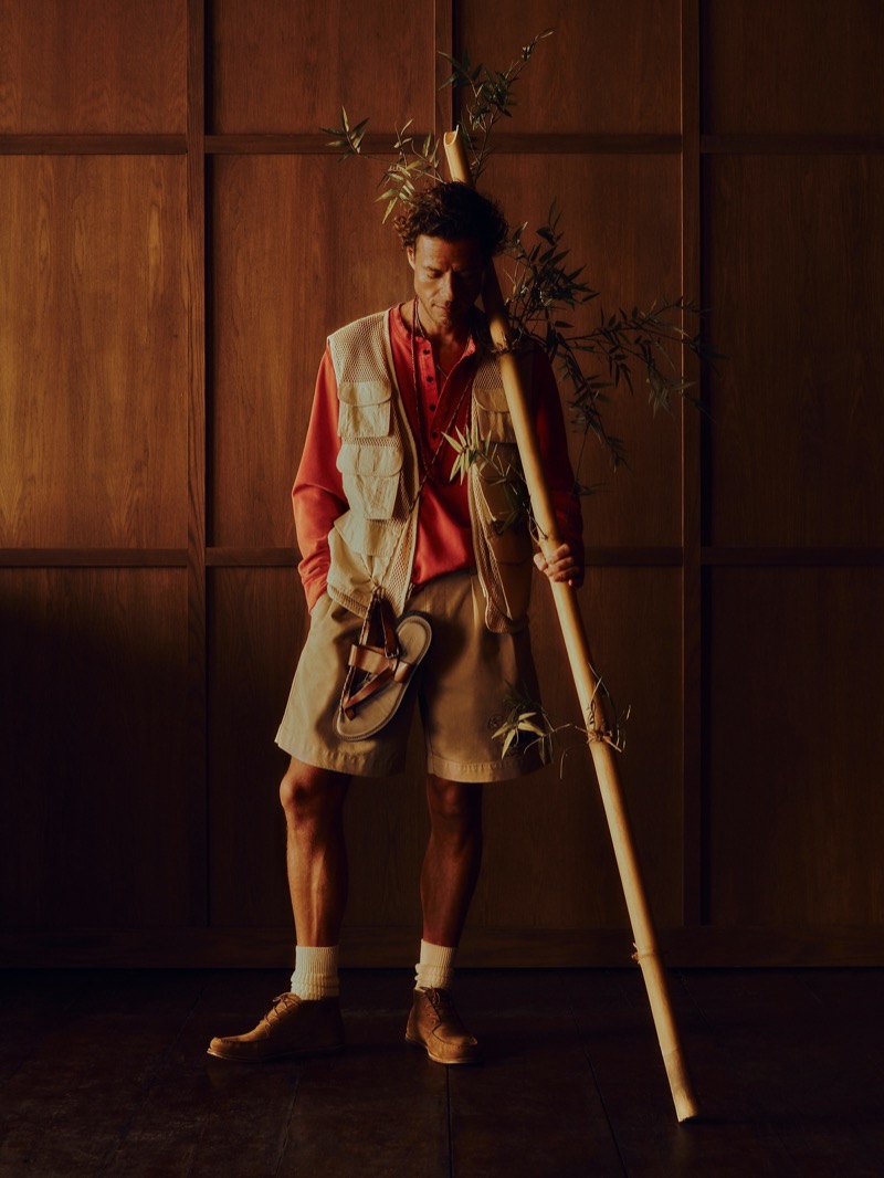 Phillips wears a multi-pocket mesh fisherman vest over a rust henley t-shirt with khaki pleated shorts, brown suede boots, and white crew socks, holding a tall bamboo pole