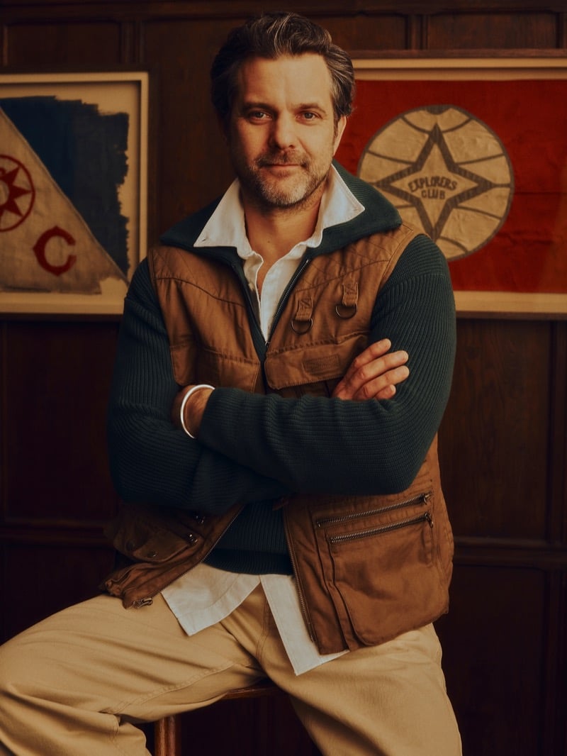 Jackson sits on a wooden stool in ribbed knitwear layered under an utility vest with a white shirt and khaki trousers, photographed in front of framed Explorers Club flags