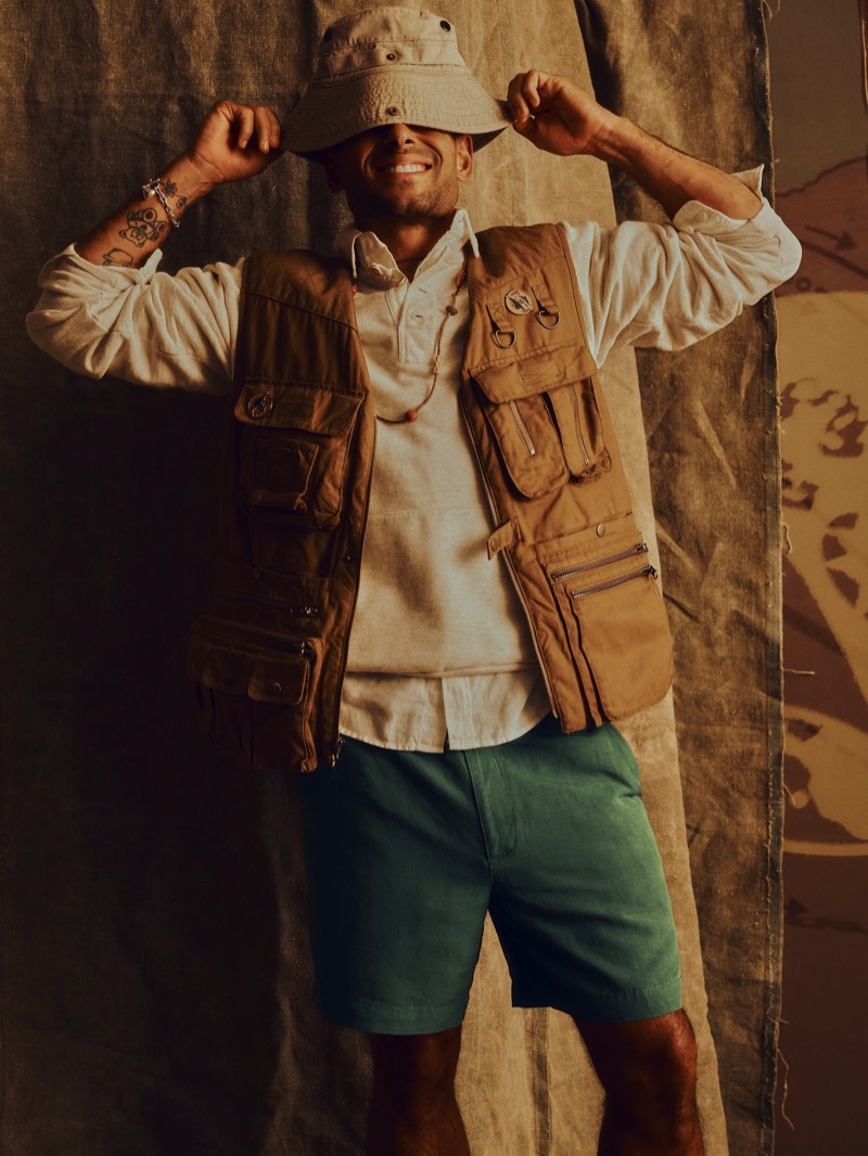 Washington wears a tan wide-brim bucket hat pulled low, a cream henley T-shirt, a tan multi-pocket photojournalist vest, and green linen shorts against a canvas backdrop for Banana Republic's Archive Reissue collection